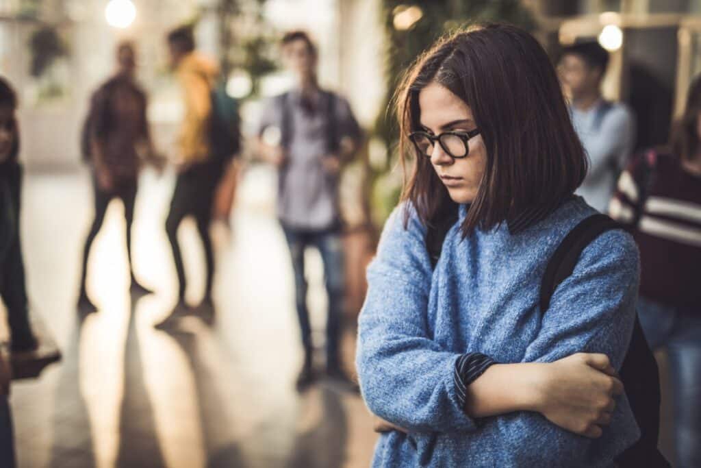 A girl wearing glasses standing off to the side from other students